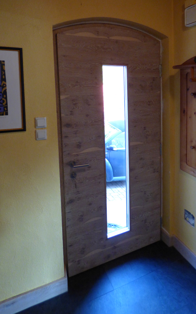 Eine hohe, schmale Holztür mit einem langen vertikalen Glasfenster, die in einer Wand mit gelbem Putz eingelassen ist, führt zu einem Außenbereich. Die Szene wird durch die warmen Farben und den dunklen Bodenbelag hervorgehoben.