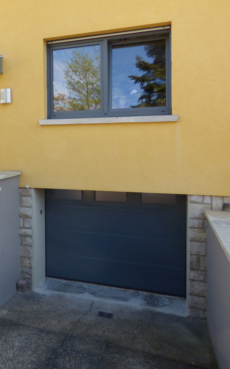 Außenansicht eines Hauses mit gelbem Putz, einem grauen Fenster mit reflektierendem Glas oben und einer dunkelblauen Garagentür unten.