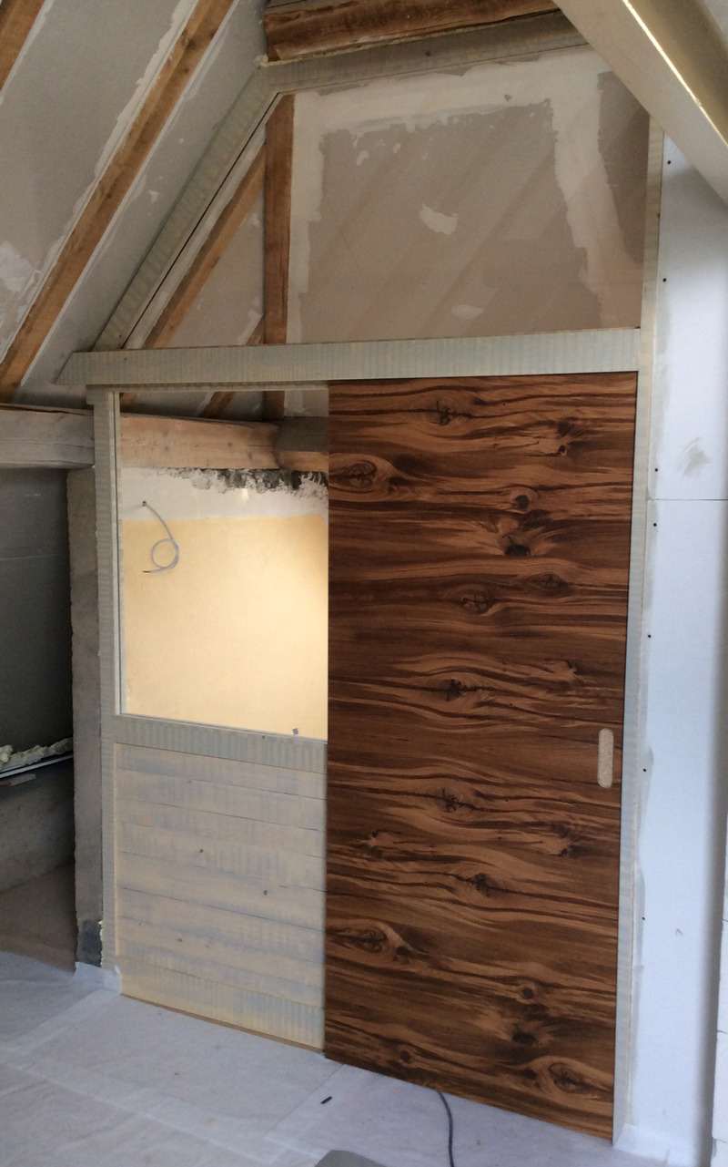 Eine Holztür mit markanter Maserung eingebaut in eine unfertige Wand mit sichtbaren Gipskartonplatten und Isoliermaterial unter einer Dachschräge.
