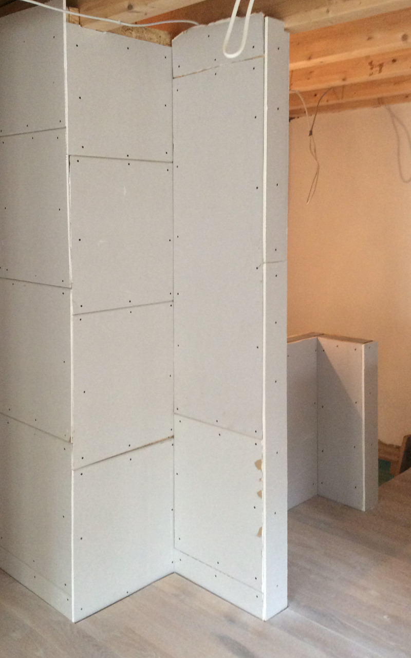 Unfertige Trockenbauwand aus Gipskartonplatten in einem Raum mit sichtbaren Holzbalken und einem Holzboden, im Prozess der Zimmerkonstruktion.