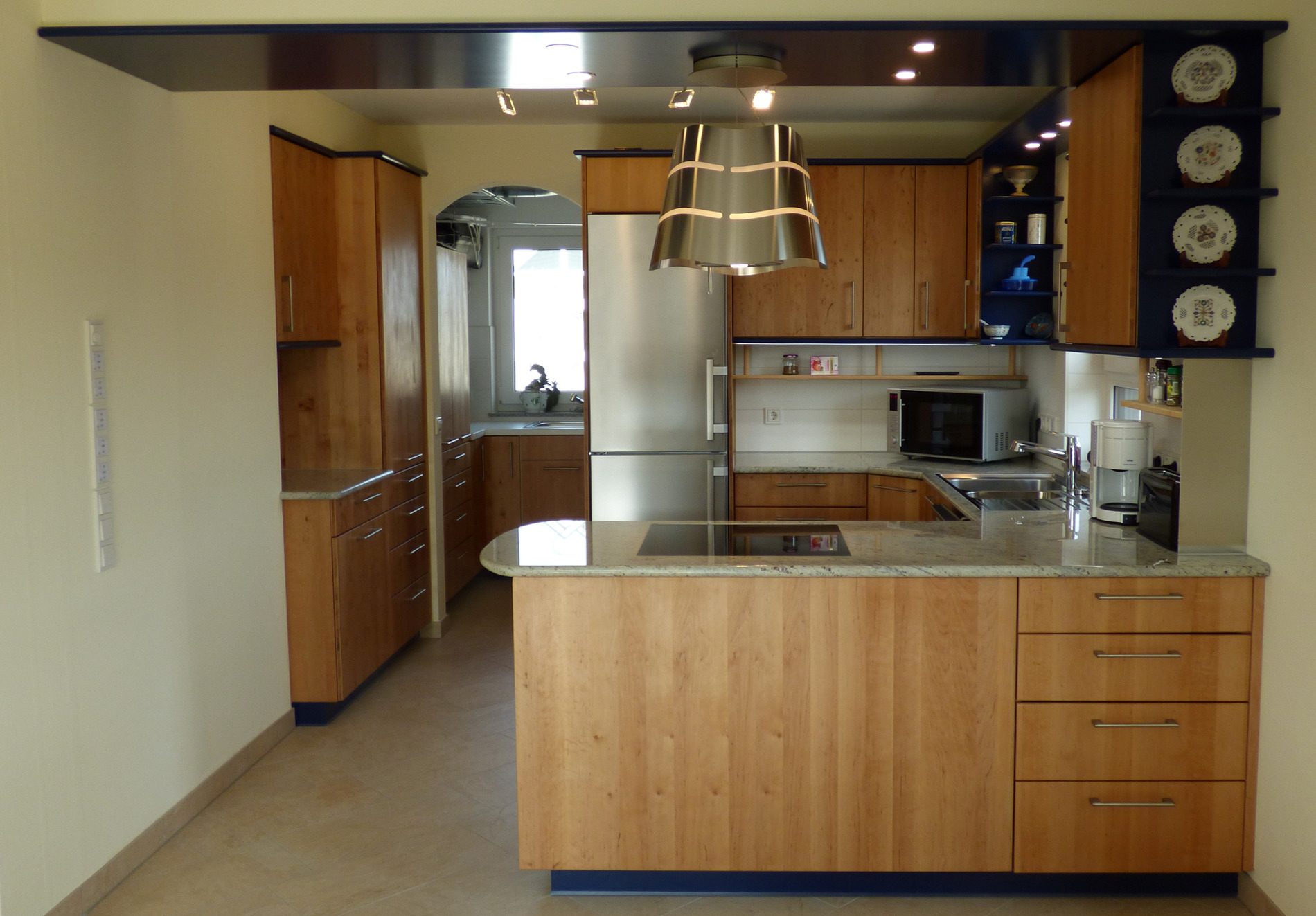 Moderne Küche mit Holzschränken, Edelstahlgeräten und einer Kochinsel mit Hängeleuchten darüber, eingebettet in ein Raumdesign mit warmen Farbtönen und dekorativen Tellern an der Wand.